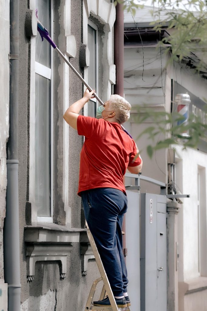 woman, elderly, standing, ladder, cleaning, window, facade, building, face mask, pandemic, covid, urban